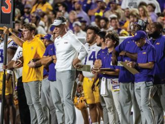 LSU football coaches and players on the sideline during a game, some holding clipboards and play sheets.