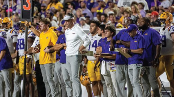LSU football coaches and players on the sideline during a game, some holding clipboards and play sheets.
