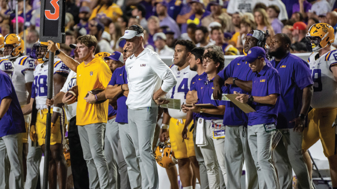 LSU football coaches and players on the sideline during a game, some holding clipboards and play sheets.
