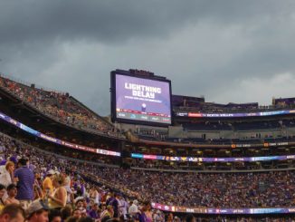 Packed football stadium with fans in purple; scoreboard reads 'Lightning Delay' during the event pause.