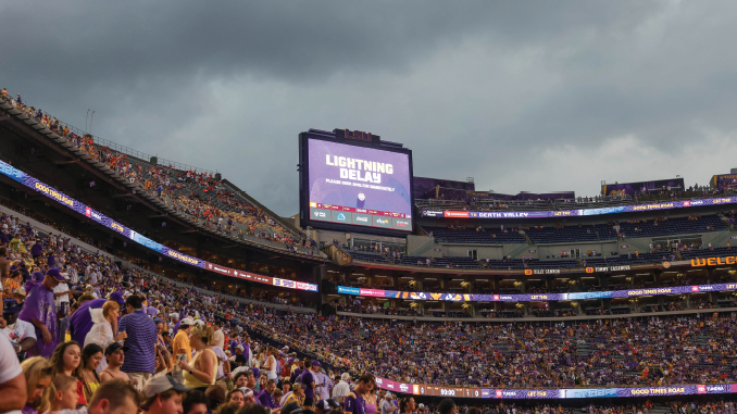 Packed football stadium with fans in purple; scoreboard reads 'Lightning Delay' during the event pause.
