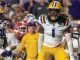LSU football player wearing number 1 in a white jersey and gold pants on the field during a game, with teammates and a referee in the background