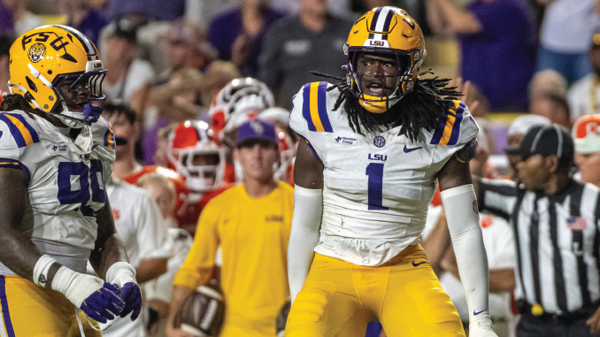 LSU football player wearing number 1 in a white jersey and gold pants on the field during a game, with teammates and a referee in the background