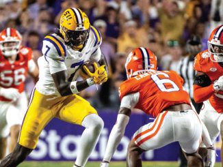 LSU running back in purple and gold carries the ball past Clemson defenders in orange uniforms during a college football game.