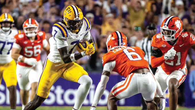 LSU running back in purple and gold carries the ball past Clemson defenders in orange uniforms during a college football game.