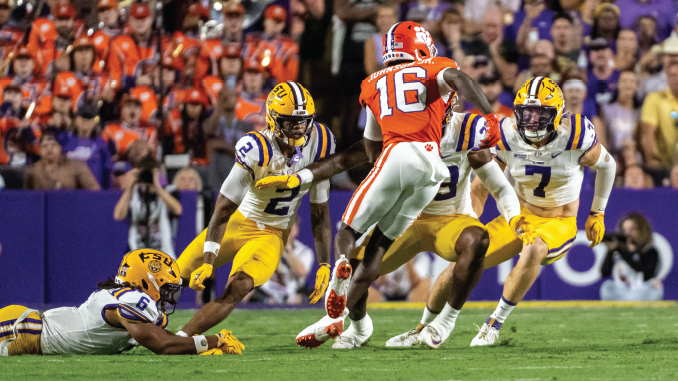 College football action: Clemson player in orange (#16) leaps over LSU defenders on the green turf with a crowded stadium in the background.