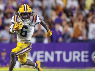 LSU running back wearing number 6 sprints with the football, pursued by Clemson defender in orange jersey on a crowded field.