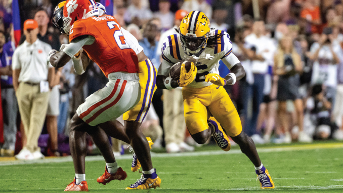 College football scene: a running back in purple-and-yellow uniform carries the ball, dodging a tackler in an orange jersey on a green field with spectators in the background.