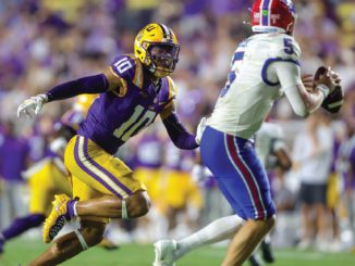 LSU defender in purple and gold (#10) chases a quarterback in white and red on a bright game day field.