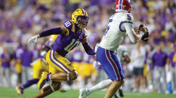 LSU defender in purple and gold (#10) chases a quarterback in white and red on a bright game day field.