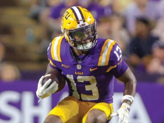 LSU football player in purple and gold jersey number 13 running with the ball on the field at a game stadium