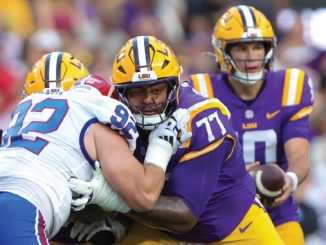 LSU players in purple and gold tackle a defender in a white jersey during a college football game on the field.