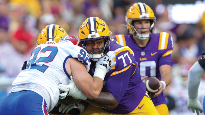 LSU players in purple and gold tackle a defender in a white jersey during a college football game on the field.