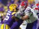 College football players collide at the line: a purple-and-gold defender (#65) tackles a white-jersey opponent with a red helmet.