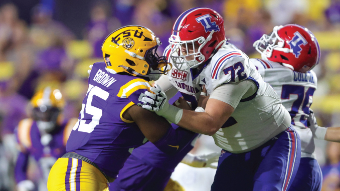 College football players collide at the line: a purple-and-gold defender (#65) tackles a white-jersey opponent with a red helmet.