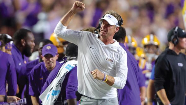 LSU football coach on the sidelines celebrating with a clenched fist amid players in purple and gold after a play era.