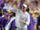 LSU football coach on the sidelines celebrating with a clenched fist amid players in purple and gold after a play era.