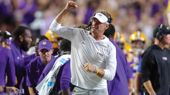 LSU football coach on the sidelines celebrating with a clenched fist amid players in purple and gold after a play era.