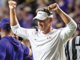 LSU football coach raises arms in celebration on the field, wearing white LSU-branded gear and headset.