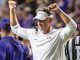 LSU football coach raises arms in celebration on the field, wearing white LSU-branded gear and headset.
