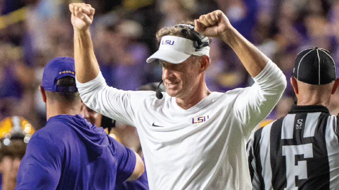 LSU football coach raises arms in celebration on the field, wearing white LSU-branded gear and headset.