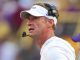 Football coach on the sideline wearing a white visor and headset, looking off to the right.