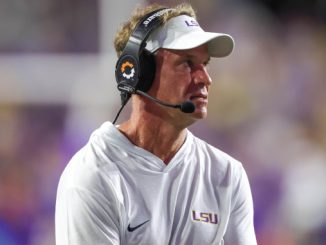 LSU football coach Lane Kiffin wearing a white polo and headset, looking toward the left on the sideline during a game.