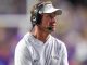 LSU football coach Lane Kiffin wearing a white polo and headset, looking toward the left on the sideline during a game.