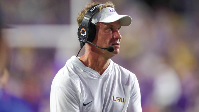 LSU football coach Lane Kiffin wearing a white polo and headset, looking toward the left on the sideline during a game.