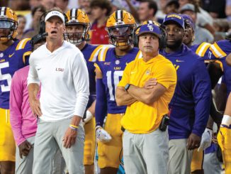 LSU football coaches and players stand on the sideline during a game, wearing purple and gold; foreground shows a coach in a white LSU polo and gray pants.