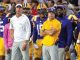 LSU football coaches and players stand on the sideline during a game, wearing purple and gold; foreground shows a coach in a white LSU polo and gray pants.