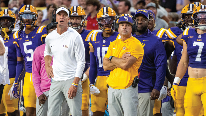 LSU football coaches and players stand on the sideline during a game, wearing purple and gold; foreground shows a coach in a white LSU polo and gray pants.