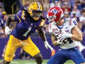 College football players on a stadium field: a purple-and-gold defender pursues a ball carrier in white with a red helmet holding the ball behind him.