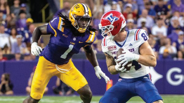 College football players on a stadium field: a purple-and-gold defender pursues a ball carrier in white with a red helmet holding the ball behind him.