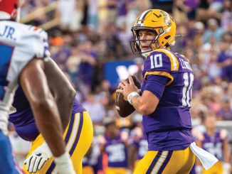 LSU quarterback in purple and gold (#10) prepares to throw a pass during a football game against the Bills, crowd in background.