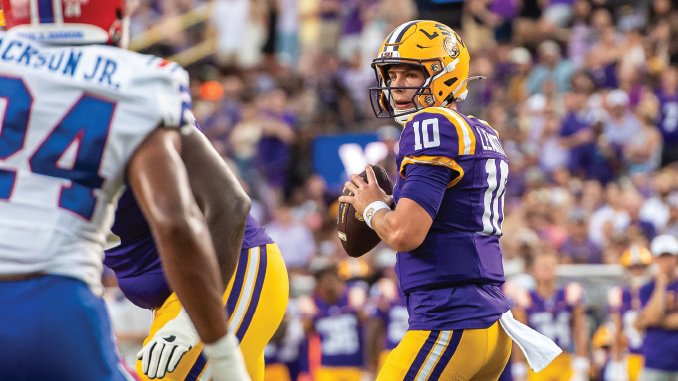 LSU quarterback in purple and gold (#10) prepares to throw a pass during a football game against the Bills, crowd in background.