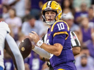 LSU quarterback in purple and gold (#10) prepares to pass during a game with a cheering crowd in the background.