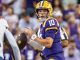 LSU quarterback in purple and gold (#10) prepares to pass during a game with a cheering crowd in the background.