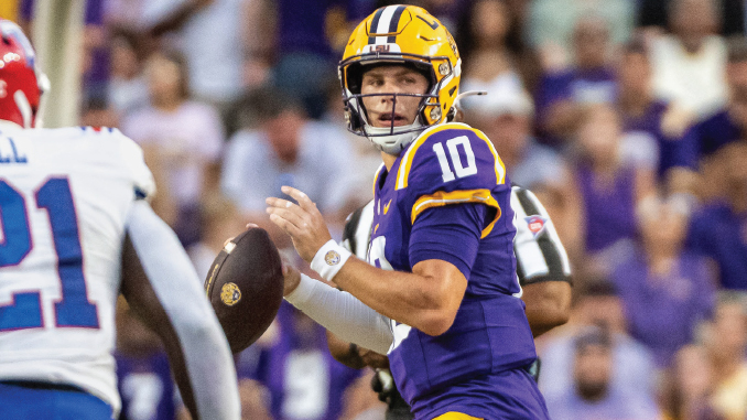 LSU quarterback in purple and gold (#10) prepares to pass during a game with a cheering crowd in the background.