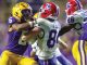 LSU defender in purple and gold tackles an Illinois player in white at the line of scrimmage during a college football game their helmets collide and players struggle for the ball.