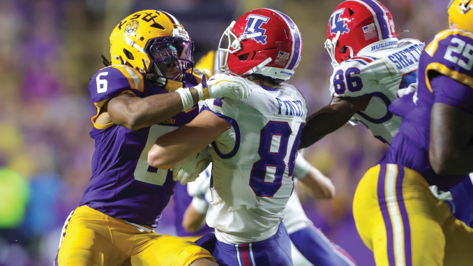 LSU defender in purple and gold tackles an Illinois player in white at the line of scrimmage during a college football game their helmets collide and players struggle for the ball.