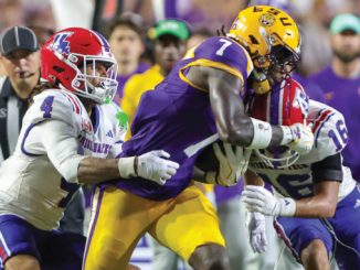LSU ball carrier in purple and gold is tackled by Florida defenders in white uniforms during a college football game on a crowded sideline.