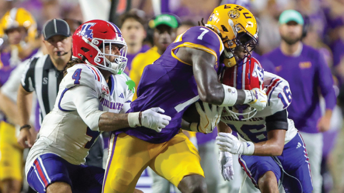 LSU ball carrier in purple and gold is tackled by Florida defenders in white uniforms during a college football game on a crowded sideline.