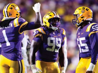 LSU football players celebrate on the field in purple and gold, giving high-fives amid a crowd.