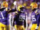 LSU football players celebrate on the field in purple and gold, giving high-fives amid a crowd.