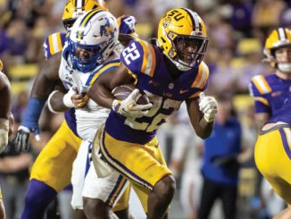 LSU Tigers running back in purple and gold carries the football as defenders chase during a college football game.