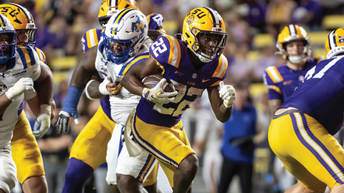 LSU Tigers running back in purple and gold carries the football as defenders chase during a college football game.