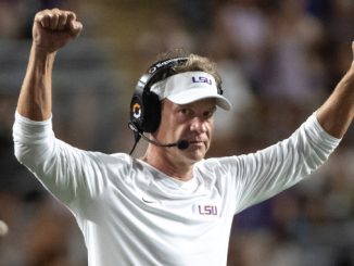 LSU football coach on the sideline, wearing a white long-sleeve shirt and visor, arms raised in celebration.