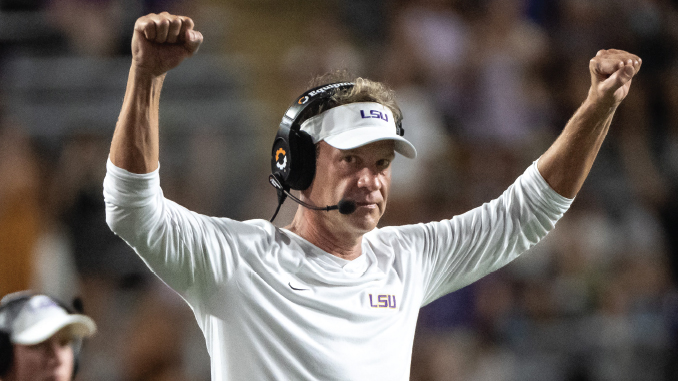 LSU football coach on the sideline, wearing a white long-sleeve shirt and visor, arms raised in celebration.
