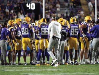 LSU football players in purple and gold snacks gathering in a huddle on the field as a coach walks through center.